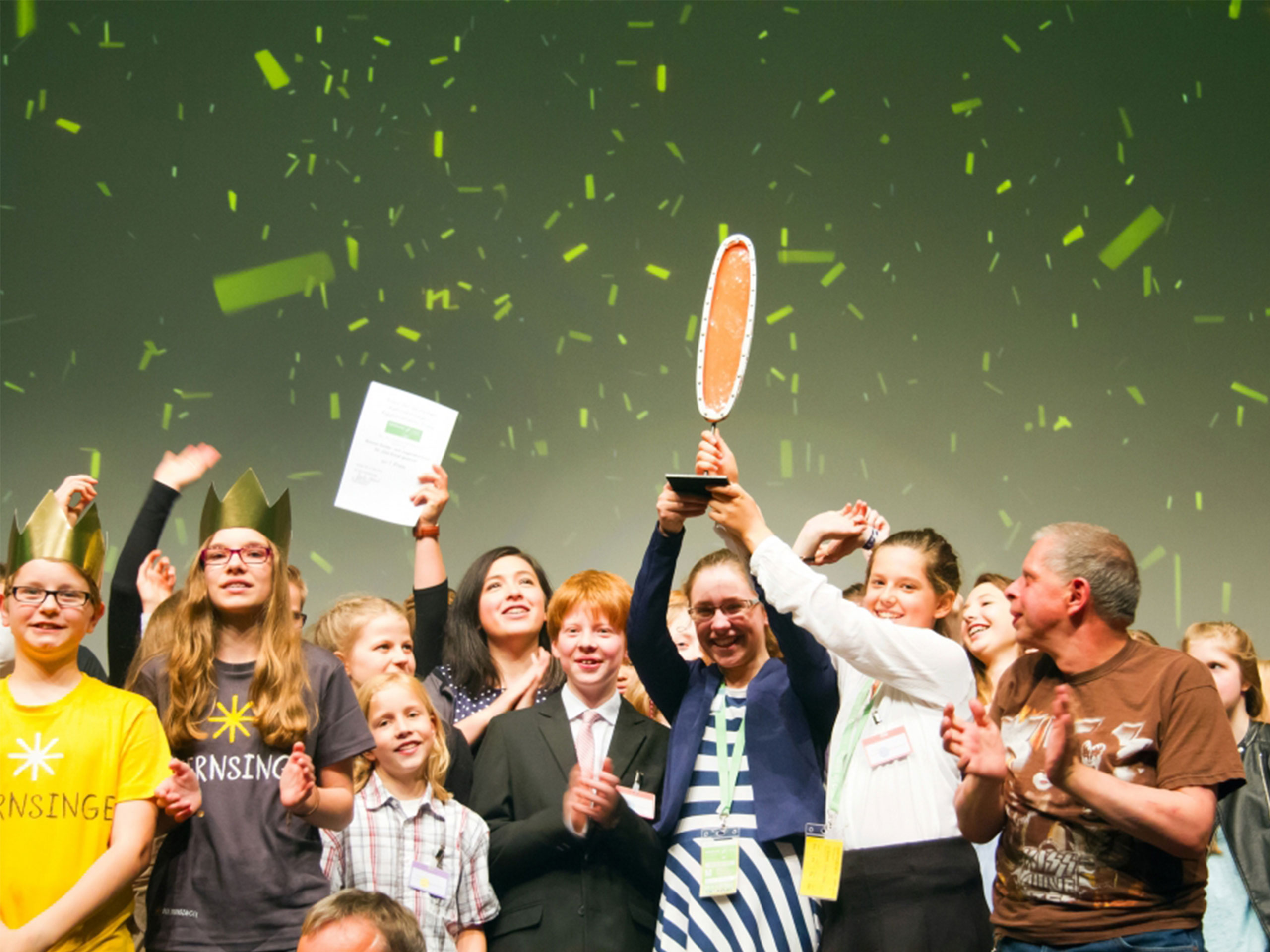 Feiernde Sternsinger-Gruppe, die den Aggiornamento-Preis gewonnen hat, und vor grüner Wand und grünem Konfetti freudestrahlend den Preis hochhalten. 